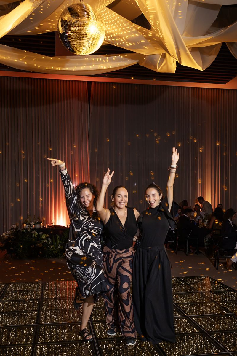 Three women pose joyfully under a disco ball at an elegant evening gala event.