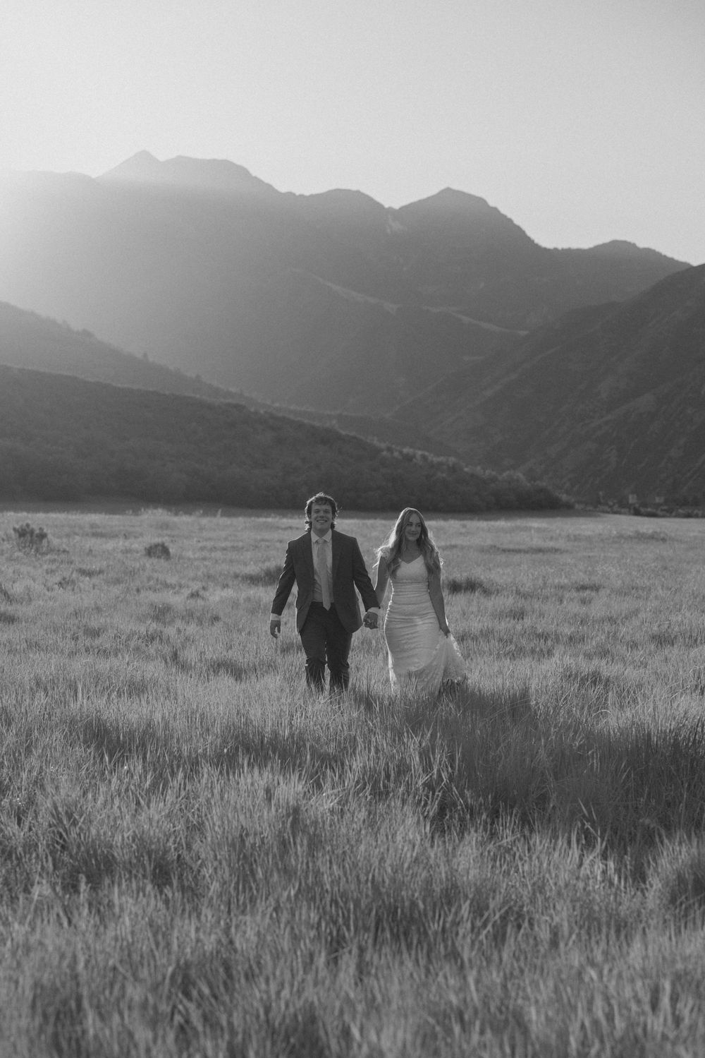 Black and white photo of a bride and groom holding hands walking through a mountain meadow at sunset.