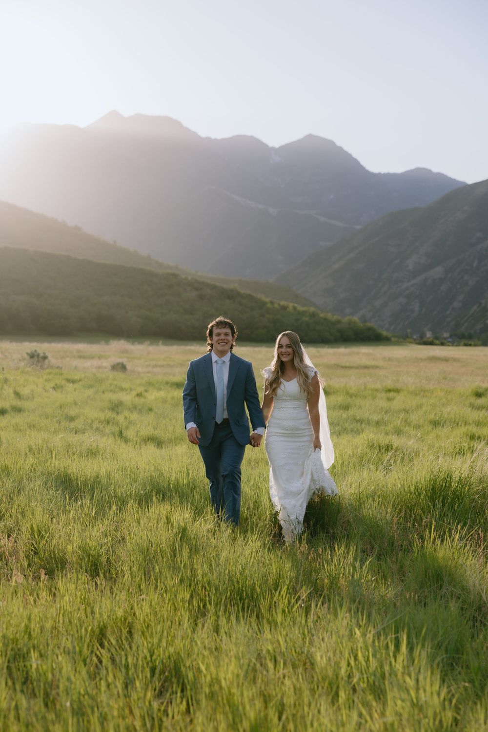 Bride and groom walk hand-in-hand through a lush green mountain meadow at golden hour.