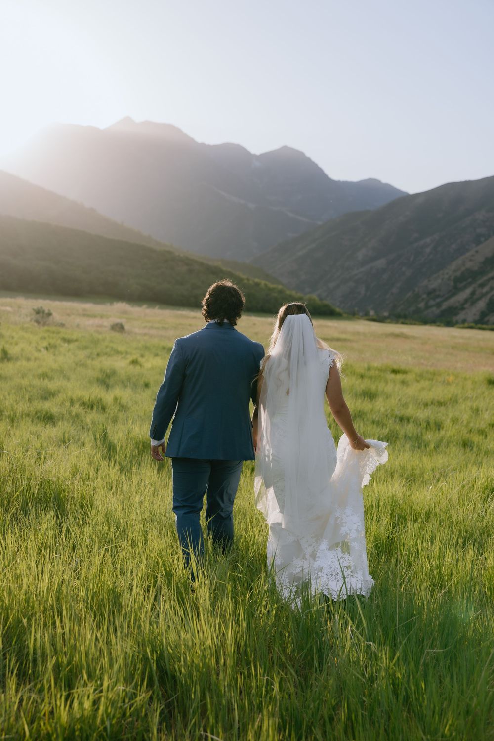 Bride and groom walking hand in hand through a lush green mountain meadow at golden hour.