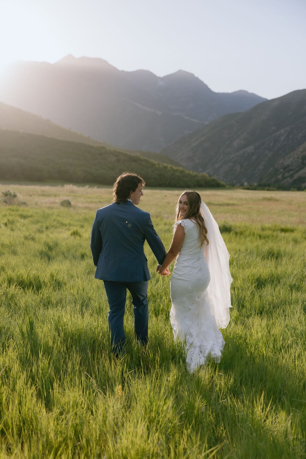 Bride and groom holding hands walking through green mountain meadow at golden hour sunset.