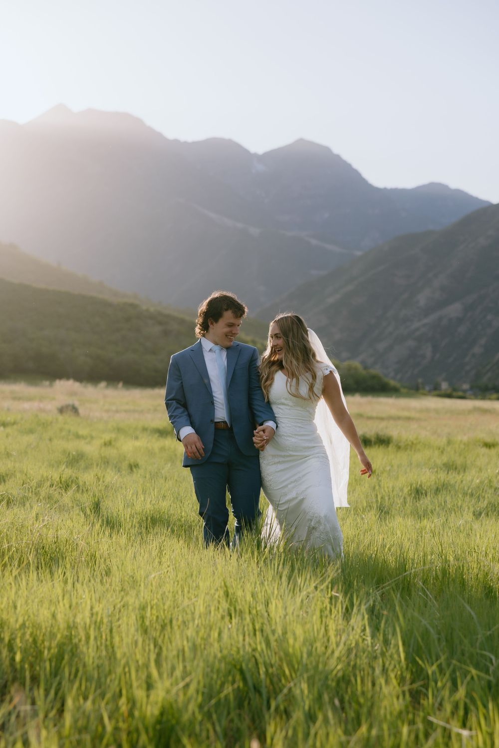 Bride and groom walking hand in hand through lush green field with stunning mountain backdrop.