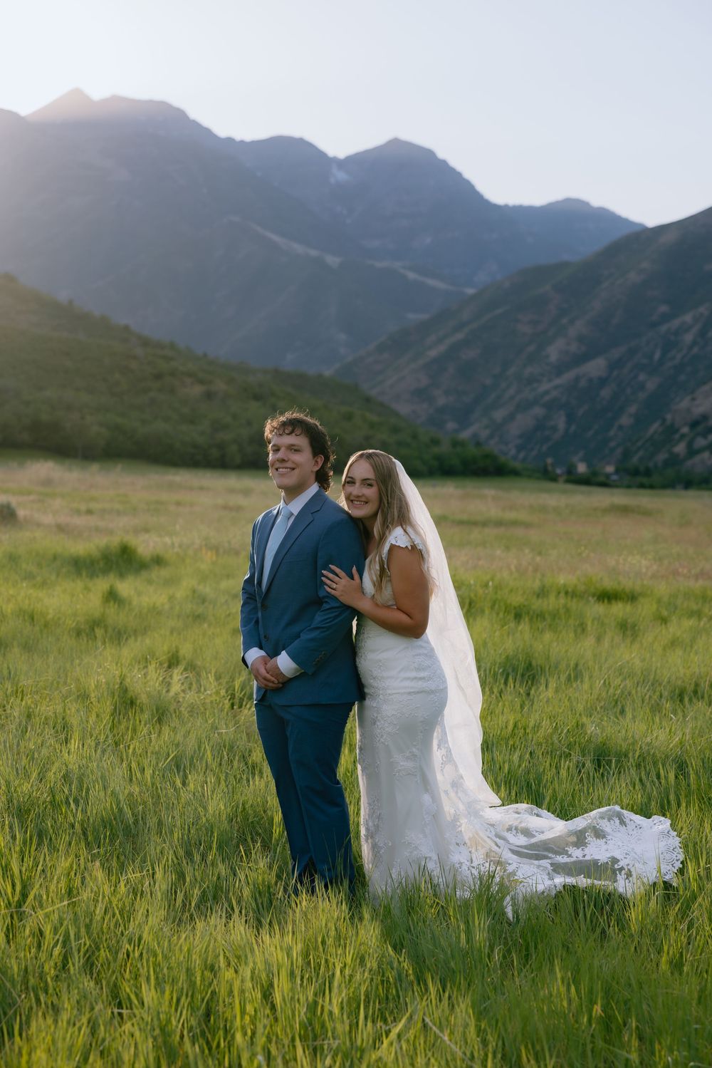 Bride and groom posing together in lush green mountain meadow at golden hour sunset.
