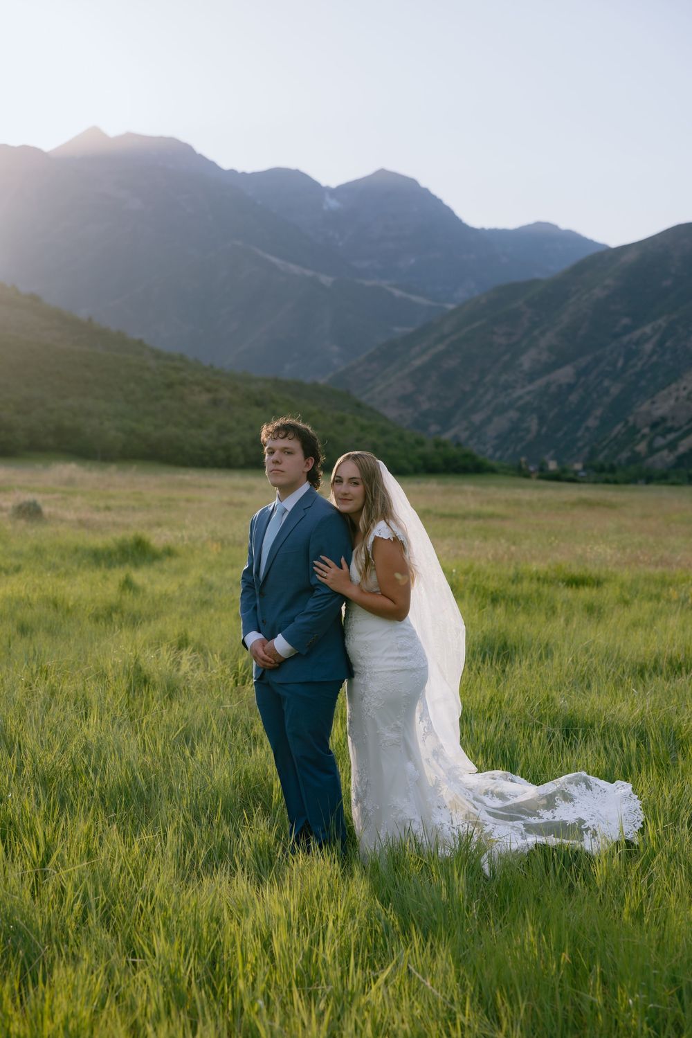 Bride in lace gown with flowing veil embraces groom in blue suit against stunning mountain backdrop.