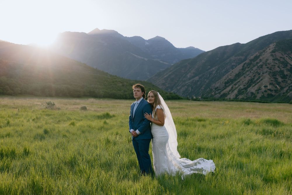 Bride and groom pose in a sunlit green field with stunning mountain backdrop on their wedding day.