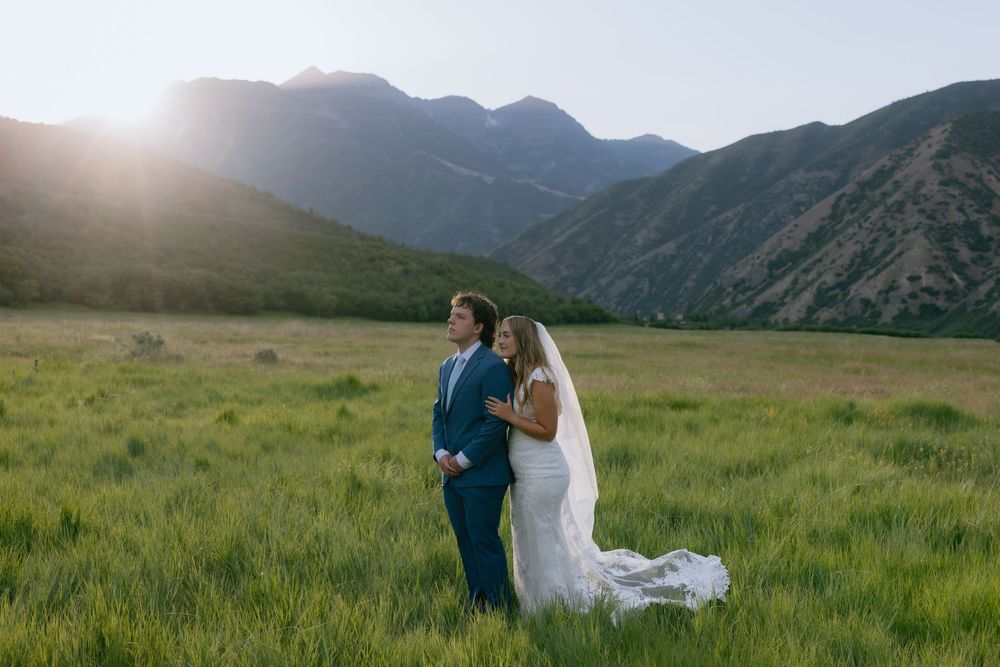 Bride and groom stand back-to-back in a green meadow with sunlit mountains in the background.