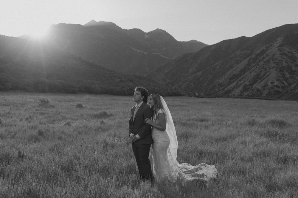 Black and white photo of bride and groom standing in a field with mountains and sunset in the background.
