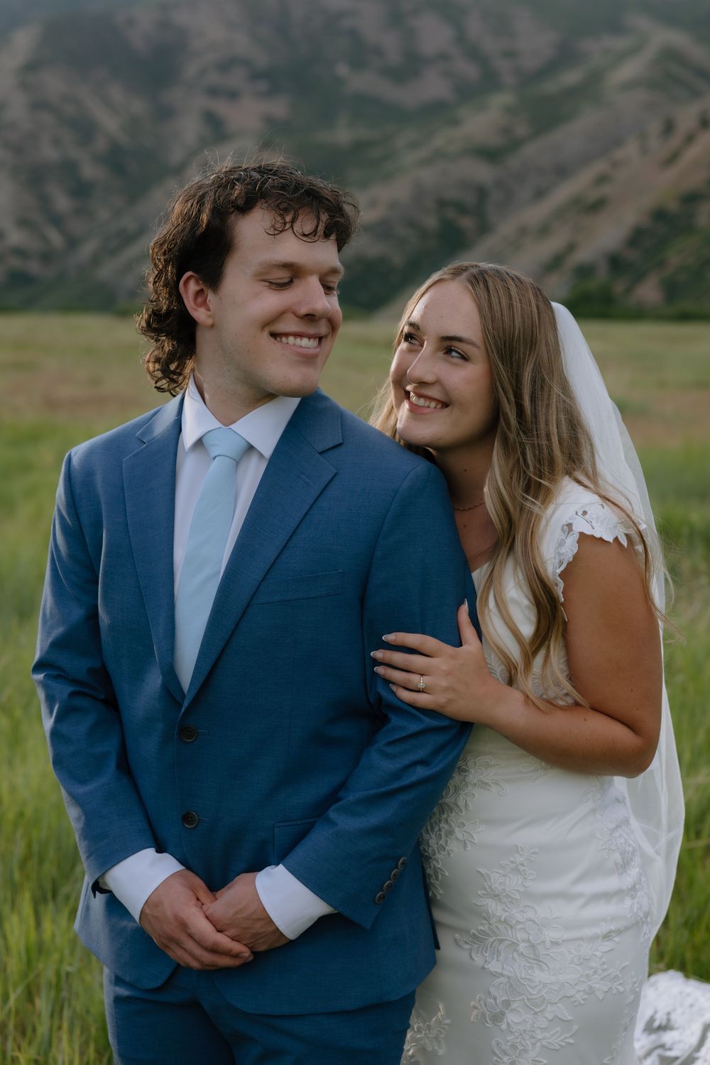 Smiling bride in lace wedding dress leans on groom in blue suit in green mountain meadow.