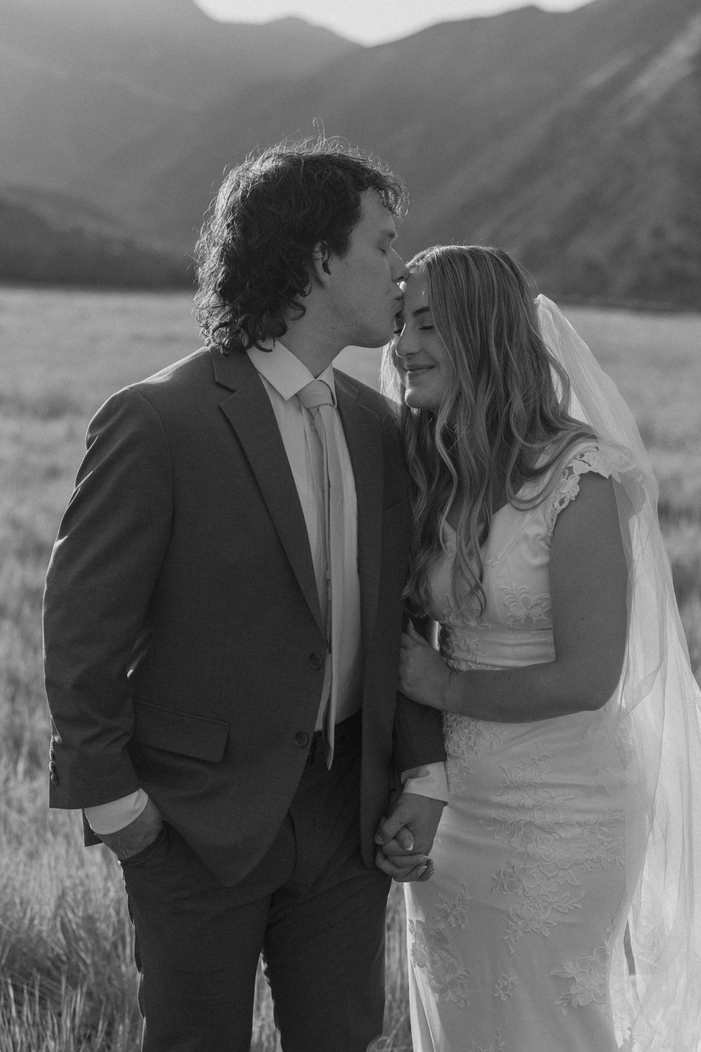 Black and white wedding photo of groom kissing bride on forehead in mountain meadow, bride wearing lace gown and veil.