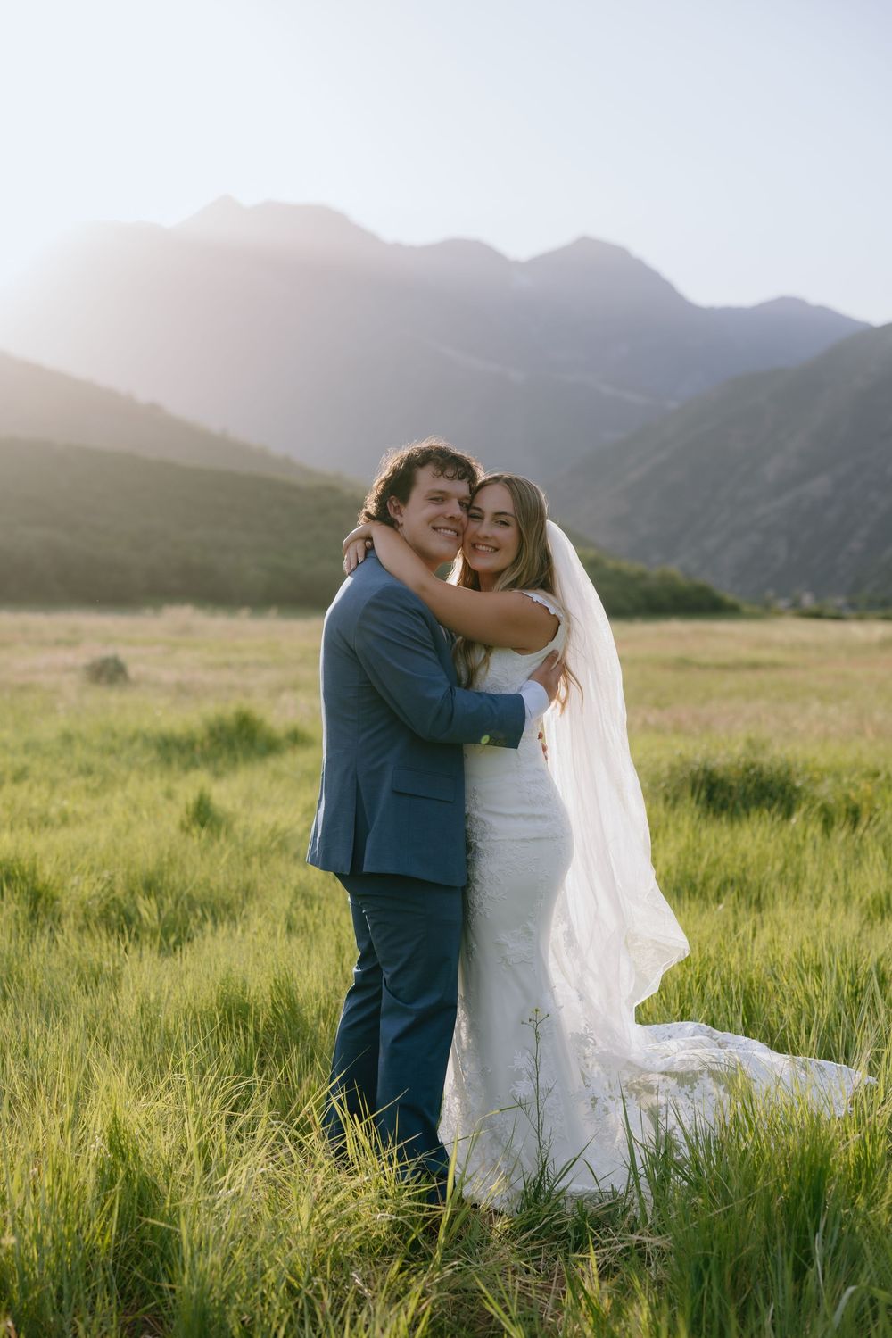 Bride and groom embracing in sunlit green mountain meadow, bride wearing lace dress and veil.