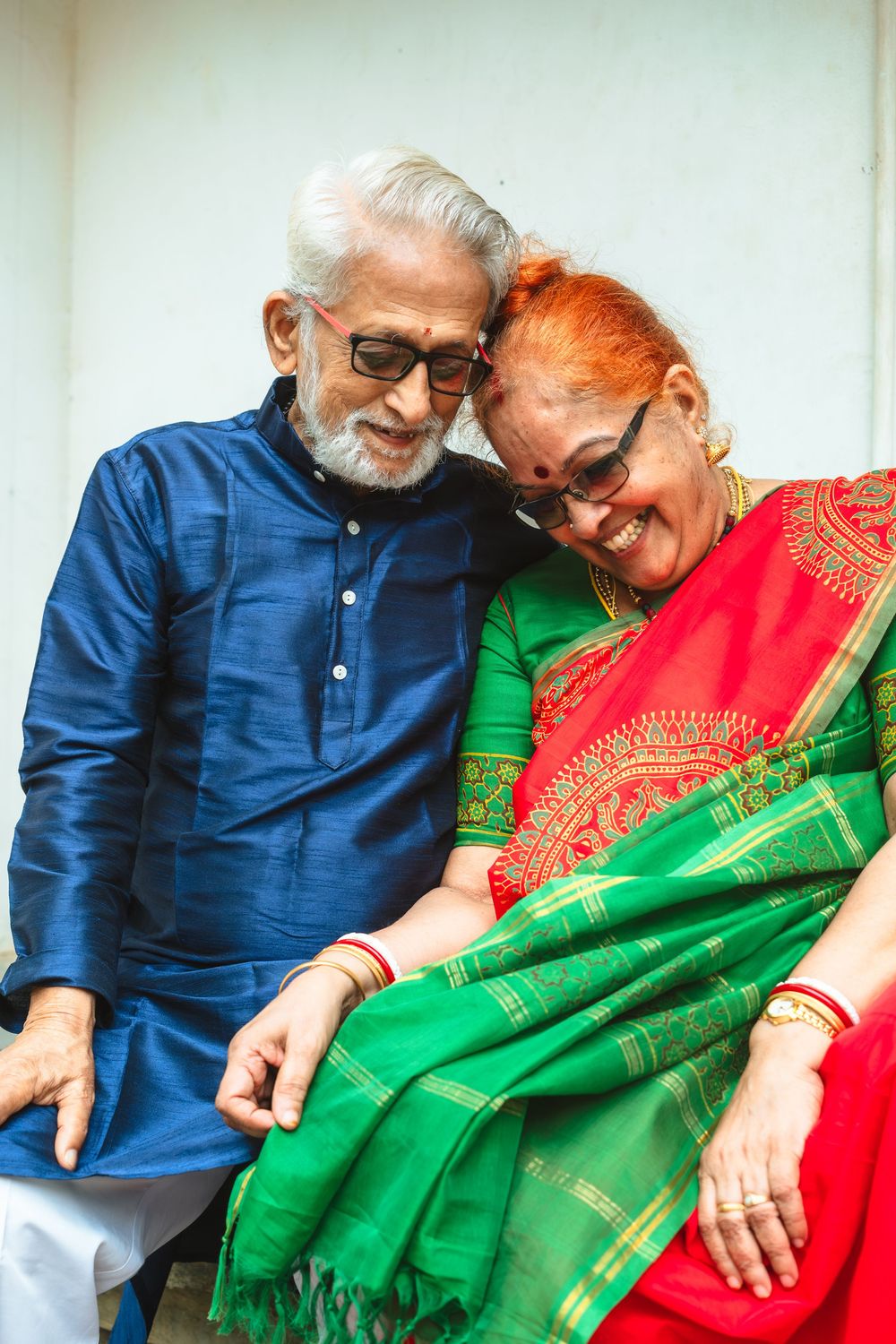 Smiling elderly couple sitting close together, the man in a blue kurta and the woman in a red and green silk saree, sharing a joyful moment.