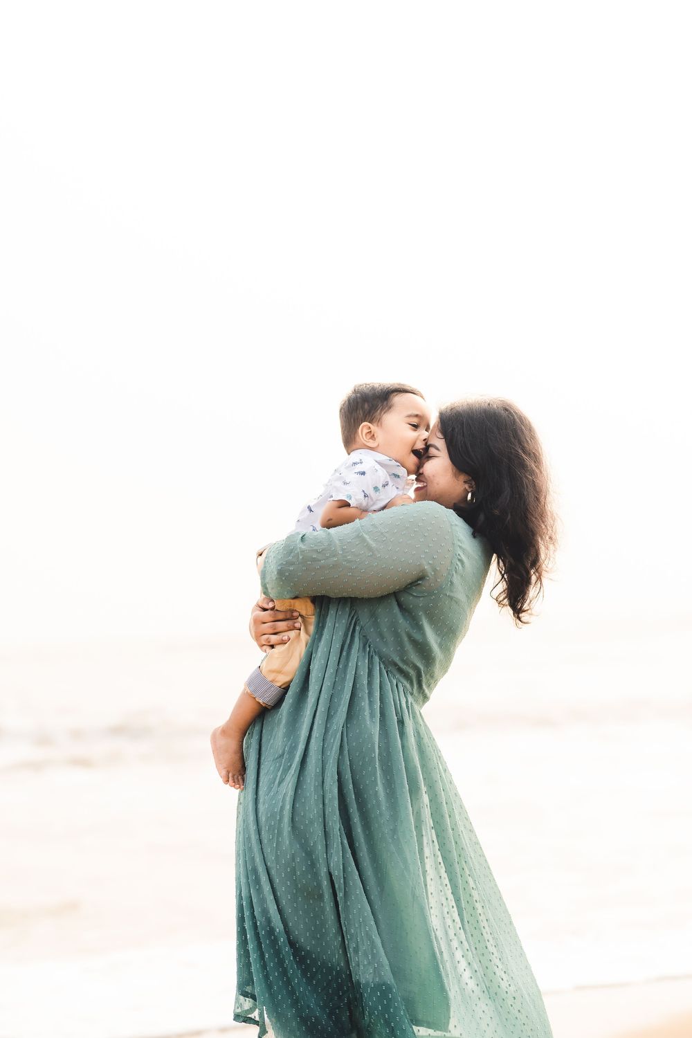A woman in a flowing green dress lovingly embraces and kisses a young child on a bright, misty beach.