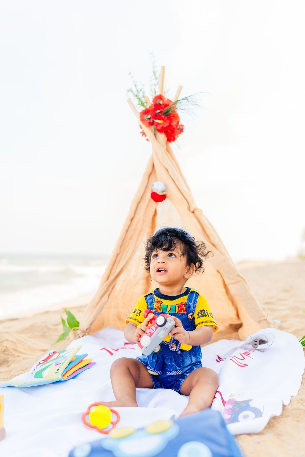 Curly-haired toddler in denim overalls plays with toys on a white blanket beside a teepee tent decorated with red flowers on a sandy beach.