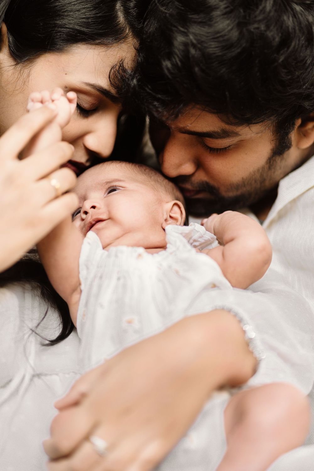 Two parents in white clothing gently kiss the forehead of their sleeping newborn baby dressed in white in a tender family portrait.