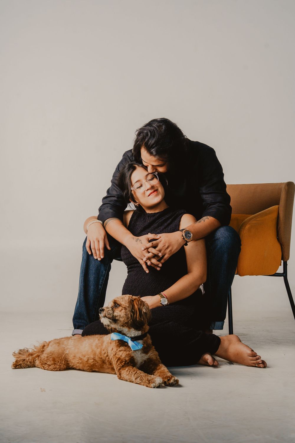 A couple in dark clothing embraces in a studio portrait, seated by an orange chair with a fluffy brown dog in a blue bow tie at their feet.