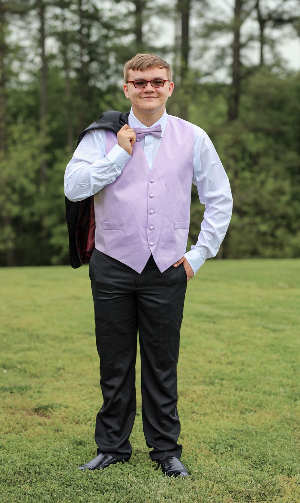 Young man in lavender vest, bow tie, and black trousers poses outdoors on grass with jacket over shoulder.