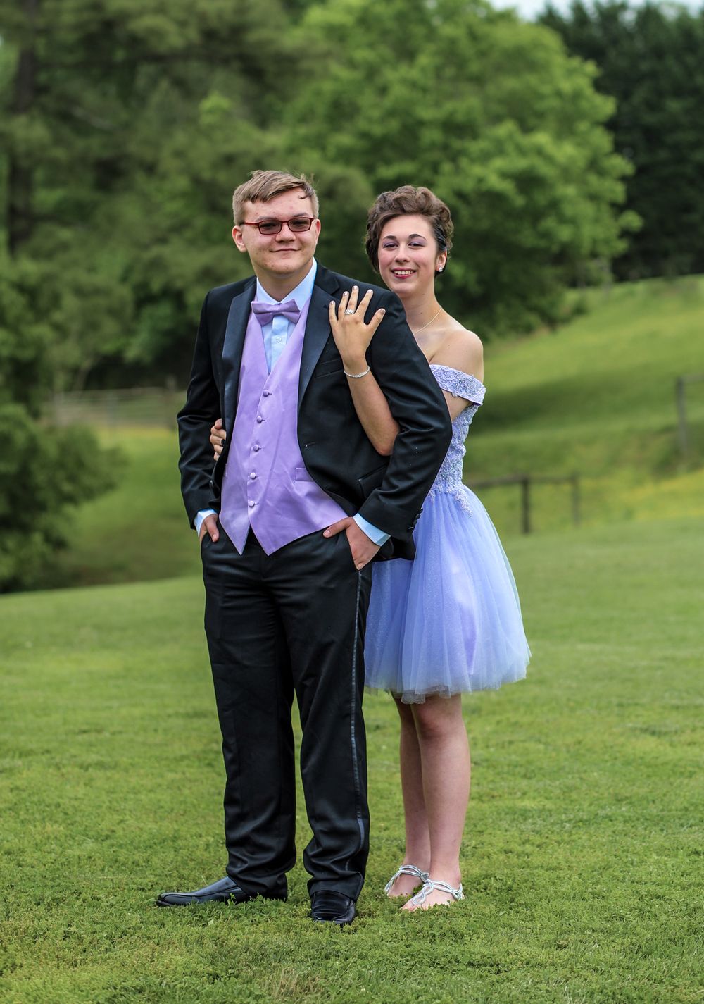 A young couple dressed in coordinating lavender prom attire poses together on a green lawn.