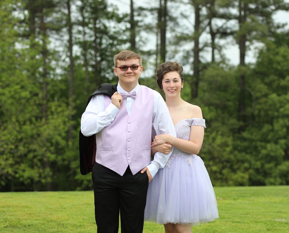 A young couple dressed in formal prom attire poses outdoors on green grass with tall trees in the background.