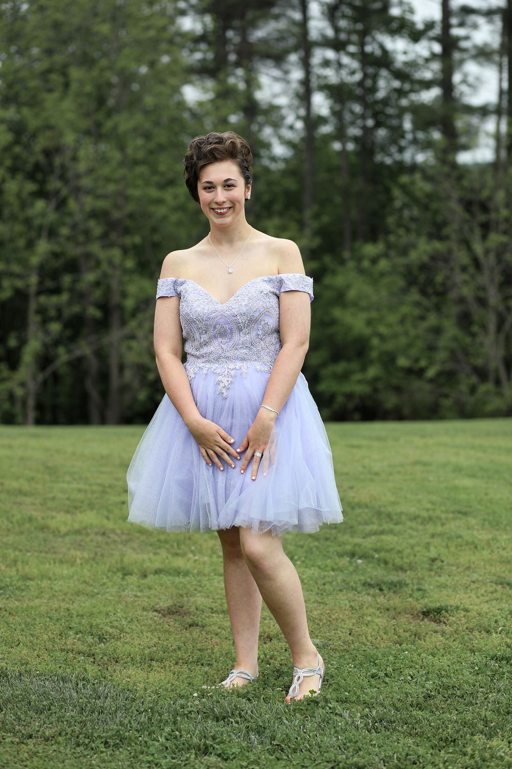 Young woman in lavender off-shoulder tulle homecoming dress posing outdoors on green grass with trees in background.