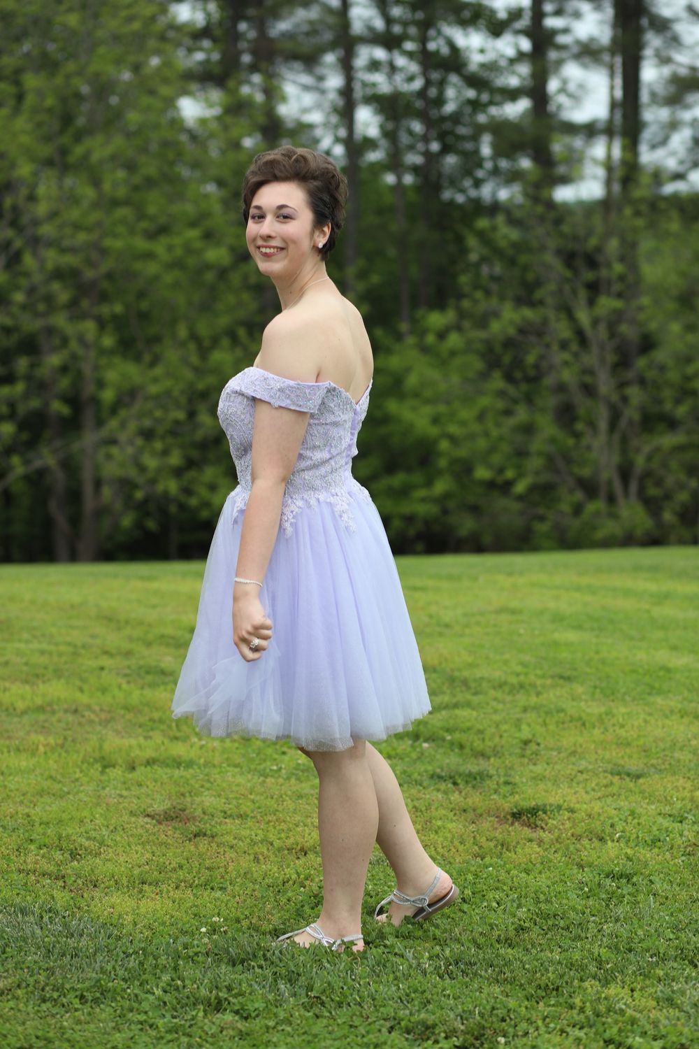 Young woman in lavender off-shoulder tulle dress posing outdoors on green grass with trees in background.