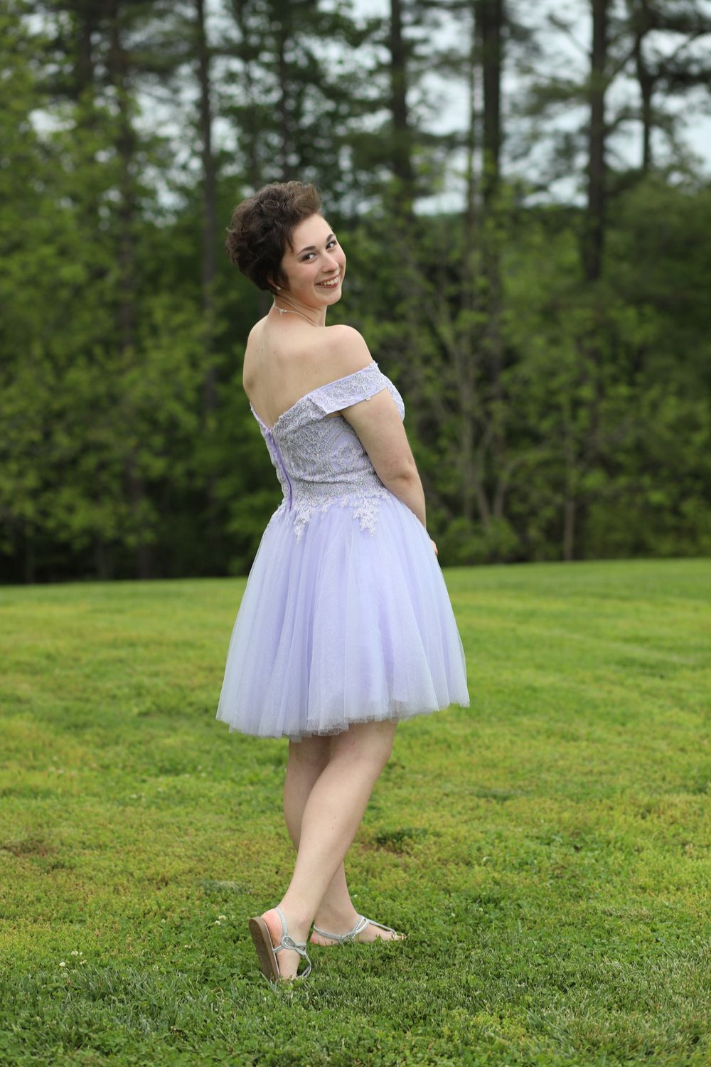 Young woman in lavender off-shoulder tulle prom dress posing outdoors on green lawn.