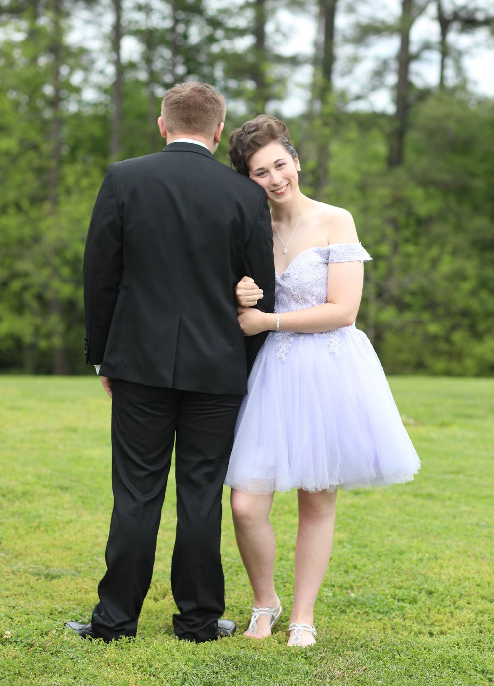 Young couple in formal attire posing outdoors, girl in lavender tulle dress smiling, boy in black tuxedo.