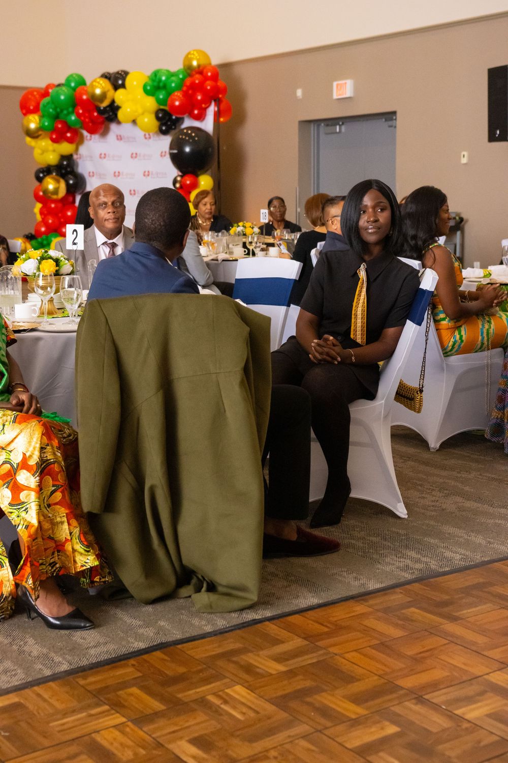 Guests in colorful African attire converse at a festive banquet hall decorated with red, yellow, green, and black balloons.