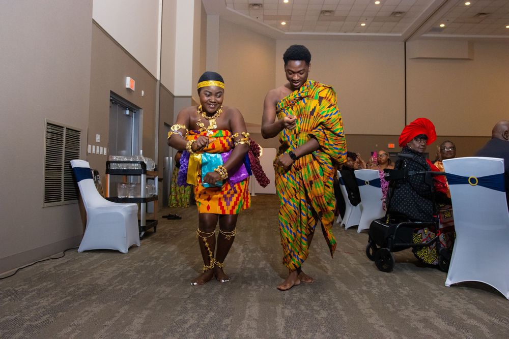 Two performers in colorful traditional Kente cloth attire dance at a cultural celebration event indoors.