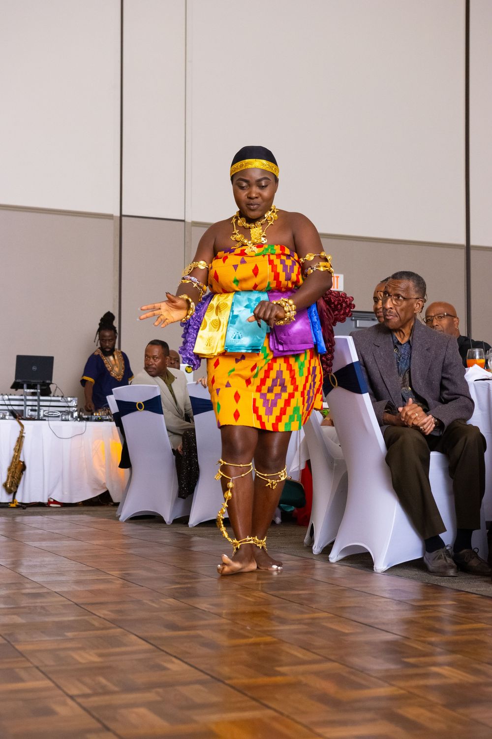 Woman in vibrant African kente dress performs traditional dance at cultural event while seated audience watches.