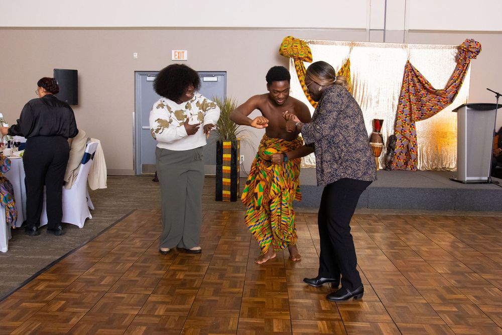 People dancing in colorful African kente cloth attire at a cultural celebration event indoors.