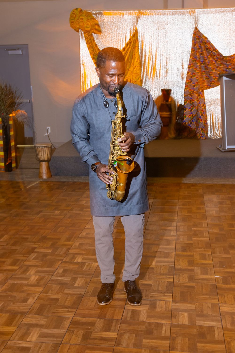 Man in gray African attire playing a gold saxophone on a wooden dance floor at an indoor cultural event.