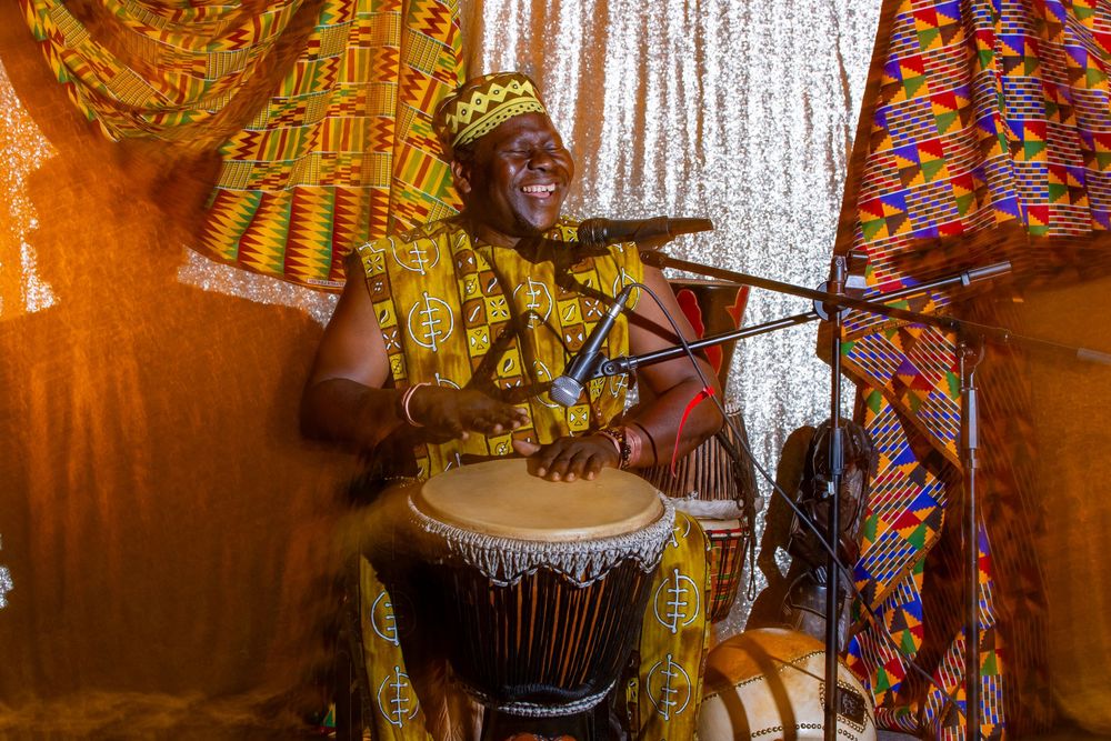 A smiling African musician in colorful kente cloth performs on a djembe drum at a vibrant cultural celebration.