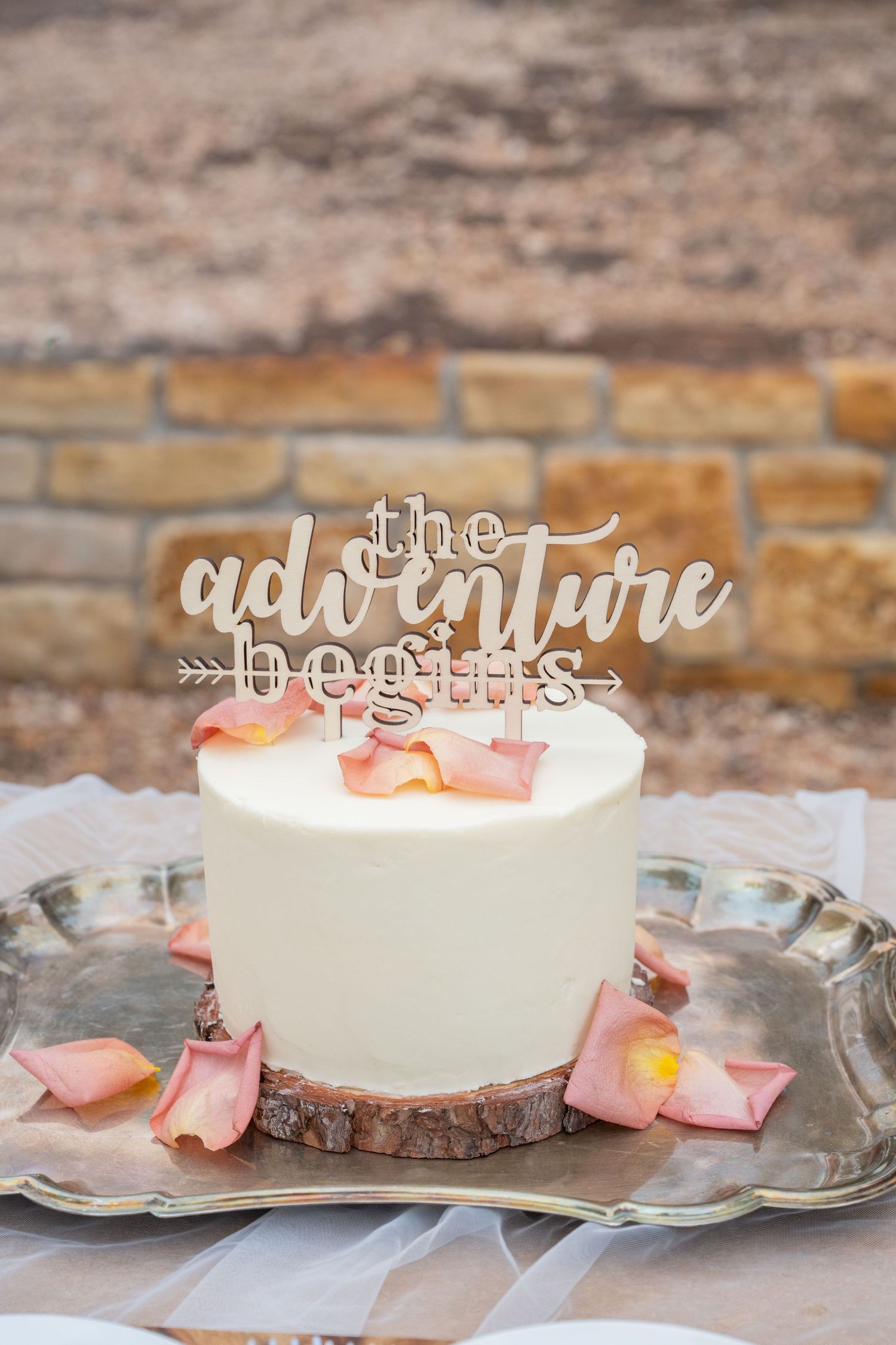 White wedding cake with 'the adventure begins' topper on wood slice, decorated with pink rose petals on silver tray.