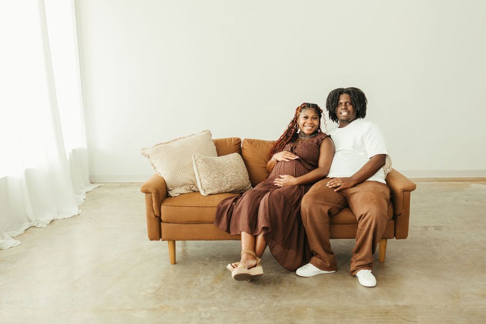 Pregnant woman in brown dress sits with partner on tan sofa during maternity photo session.