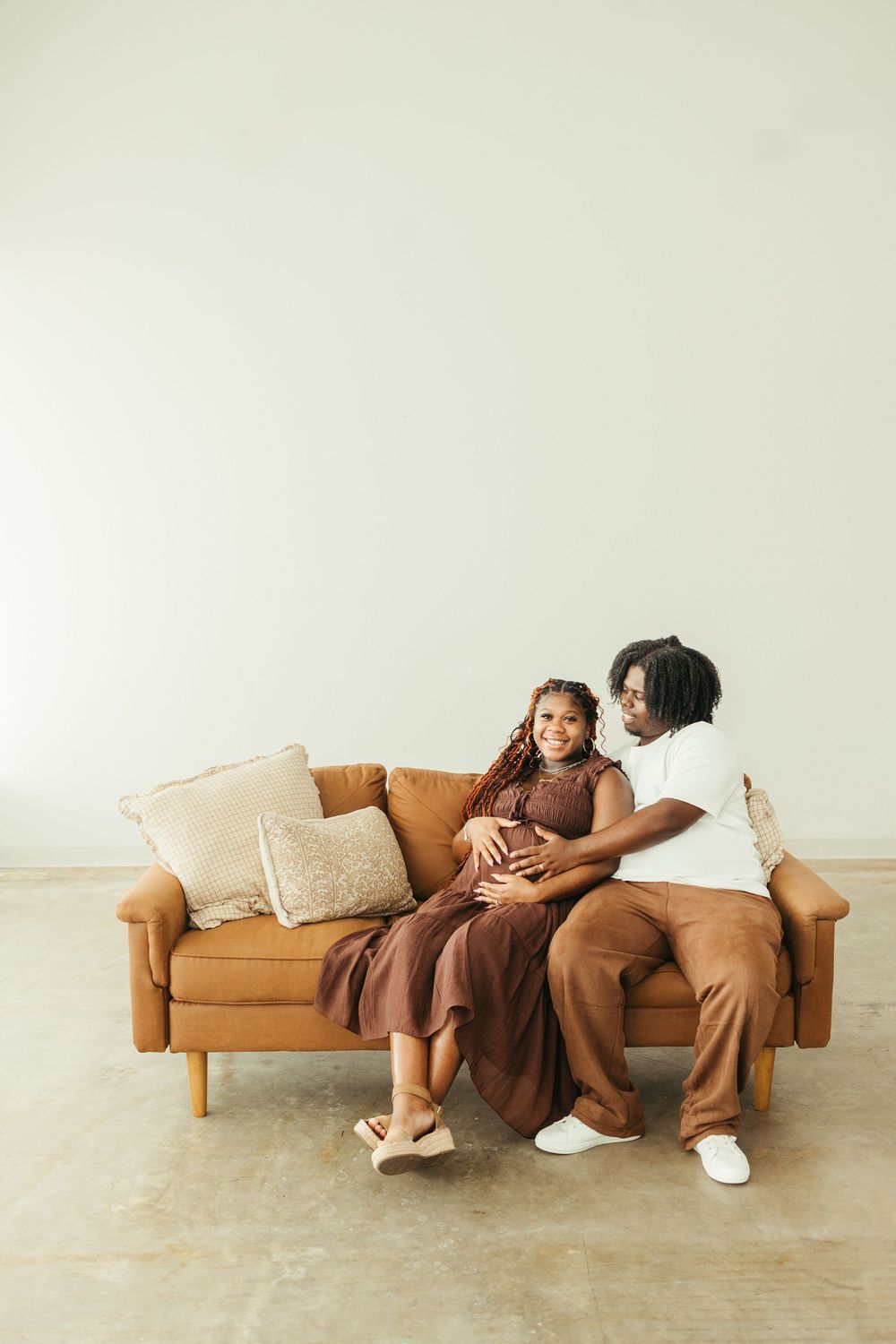 Pregnant Black couple sitting on a tan leather couch, smiling at each other while cradling her baby bump.