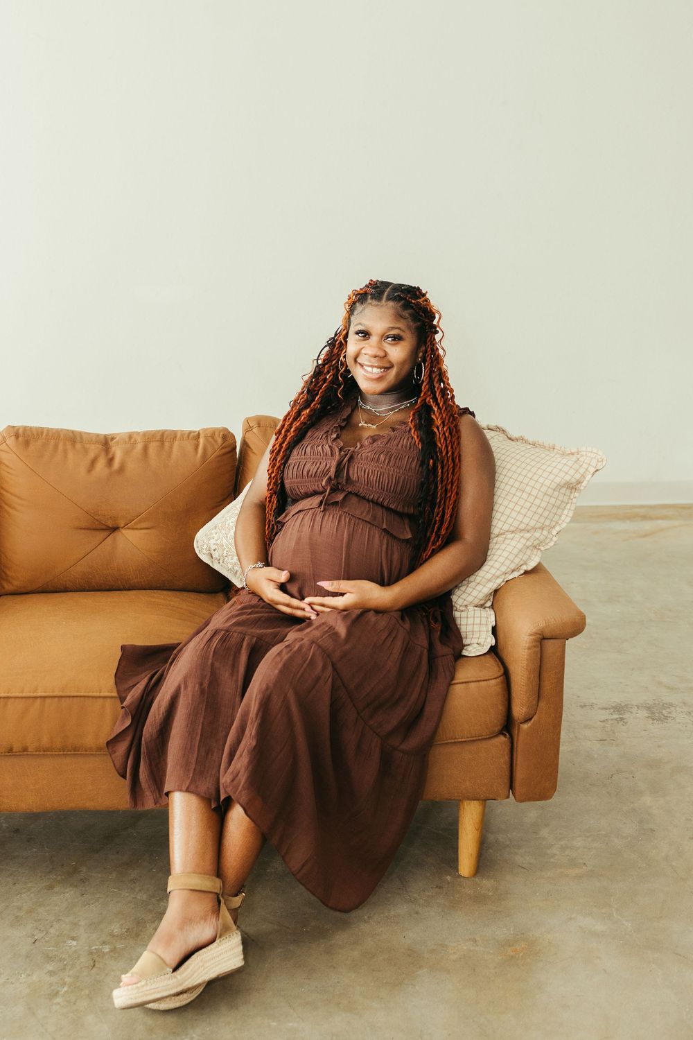 Smiling pregnant woman with braids in brown dress sits on tan sofa, hands on belly, against light gray wall.