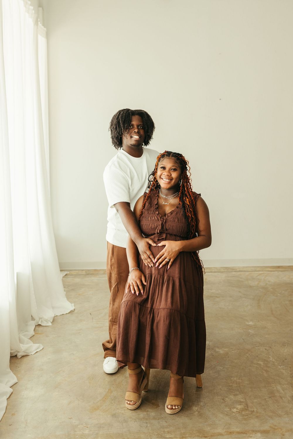 A couple poses together, the woman in a brown dress with a baby bump, both smiling in a bright studio setting.