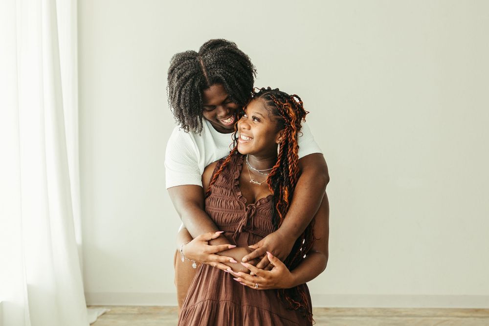 A loving Black couple embraces and smiles together in a bright white room, the woman wearing a brown dress.