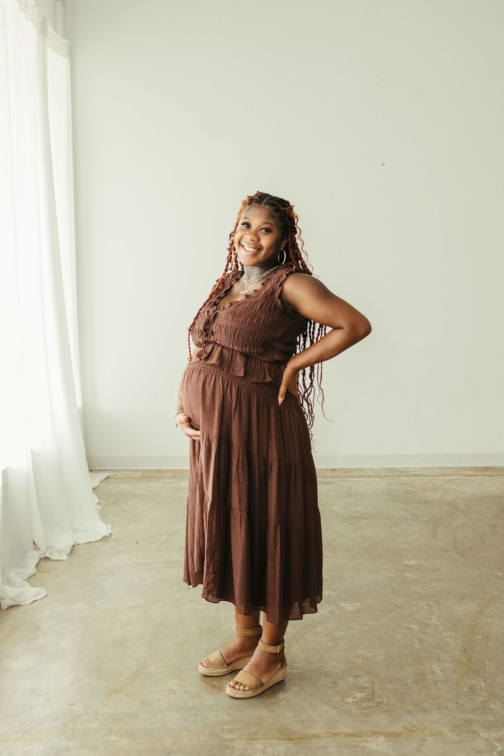 Smiling pregnant Black woman in brown tiered midi dress with braids poses in bright studio.