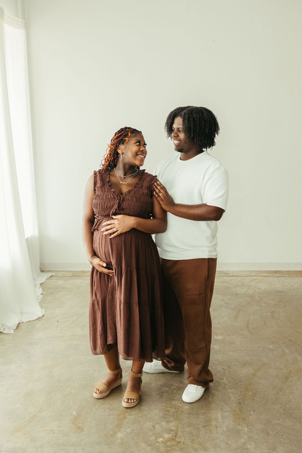 A smiling pregnant Black woman in a brown dress stands with her partner in a bright studio during a maternity photoshoot.