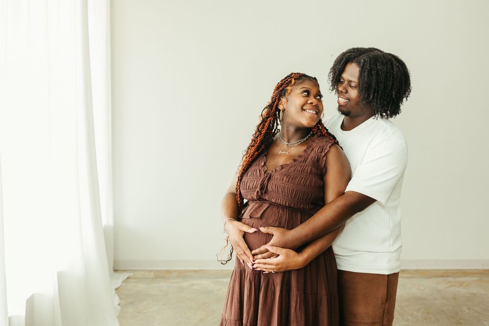 A pregnant couple smiling and embracing, with hands on baby bump, standing near a bright window.
