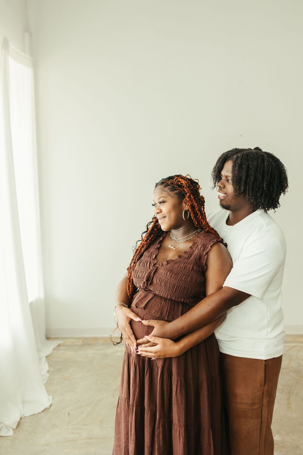 Couple embracing pregnant woman's belly in bright studio, man in white tee and brown pants.