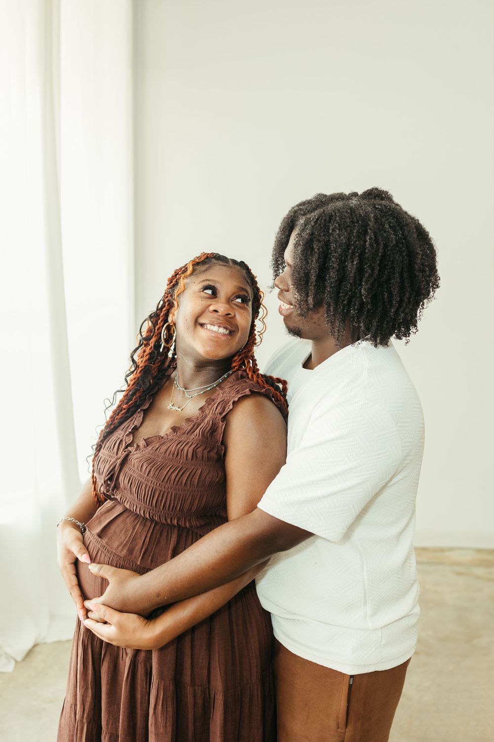 A smiling pregnant woman in a brown dress embraces her partner in a bright, minimalist room.