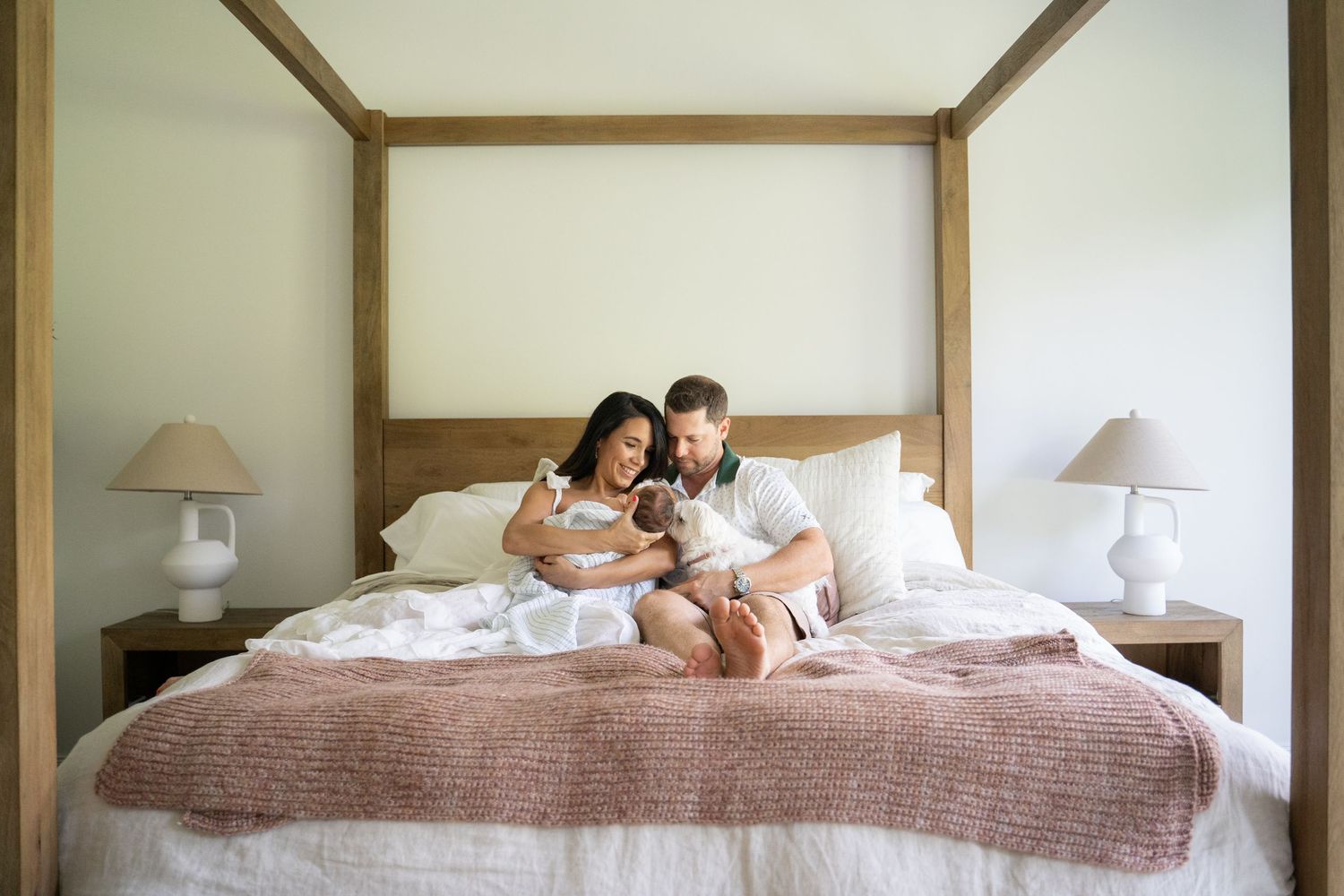 Couple sitting on a four-poster bed with a newborn baby, enjoying a peaceful moment in a bright bedroom.