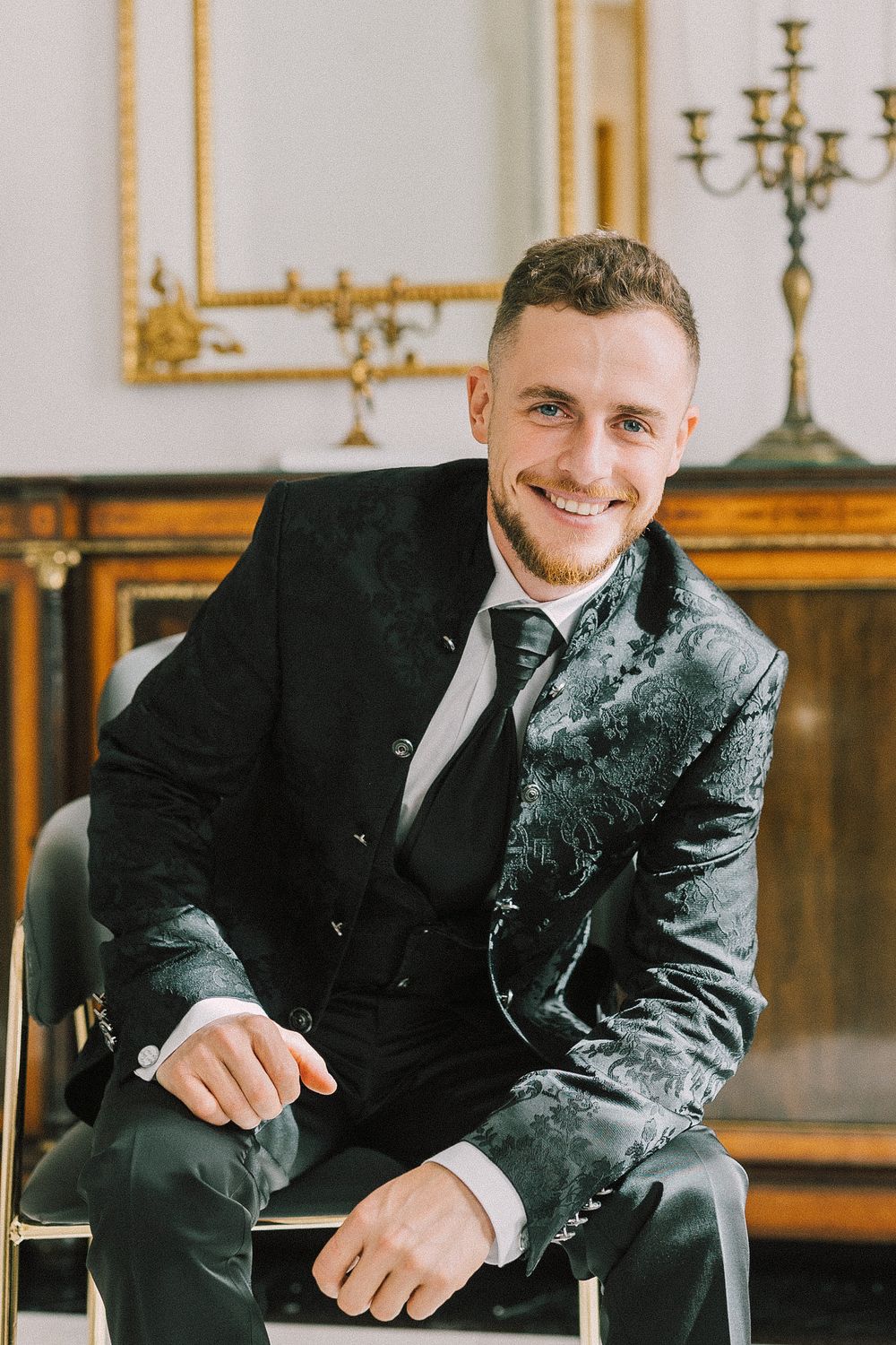 Smiling man in elegant dark velvet suit sitting on chair in ornate room with gold mirror and candelabra.