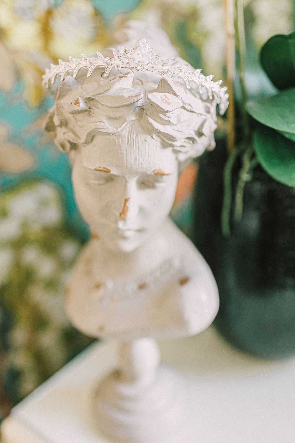 White plaster bust sculpture with floral crown and gold accents displayed on a pedestal with green foliage in background.