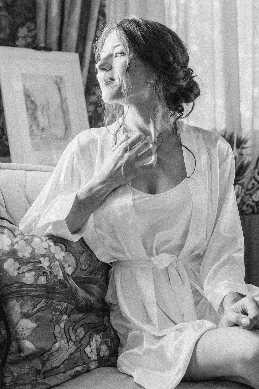 Black and white photo of a woman in a white silk robe sitting gracefully on a floral sofa near a window.