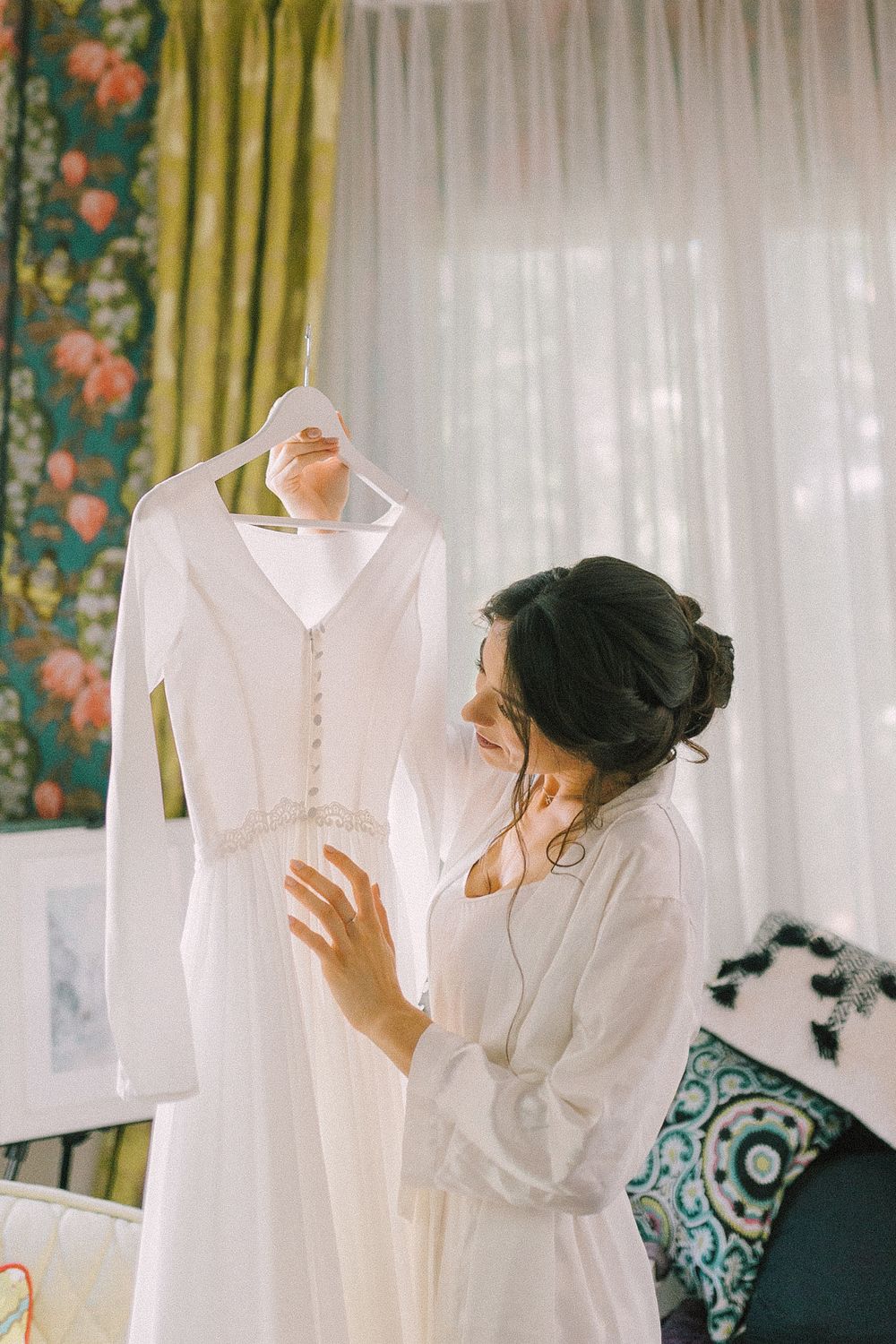 Bride in white robe admiring her elegant wedding dress on a hanger in a beautifully decorated room.