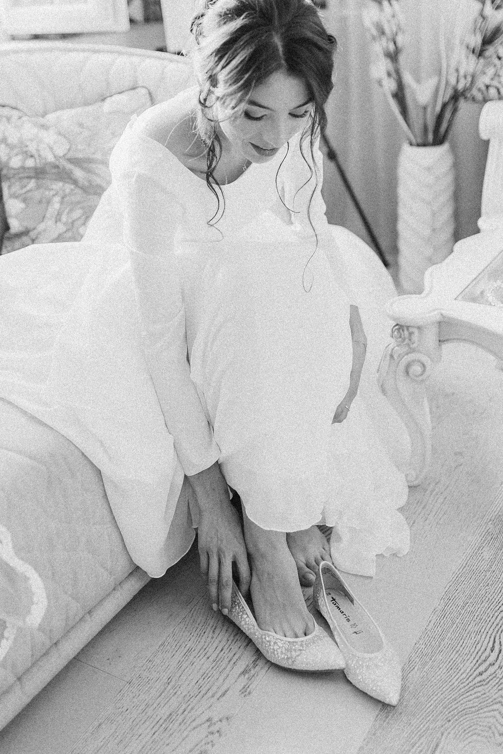 Bride in white dress putting on sparkly pointed-toe flats while seated on elegant chair in black and white photo.