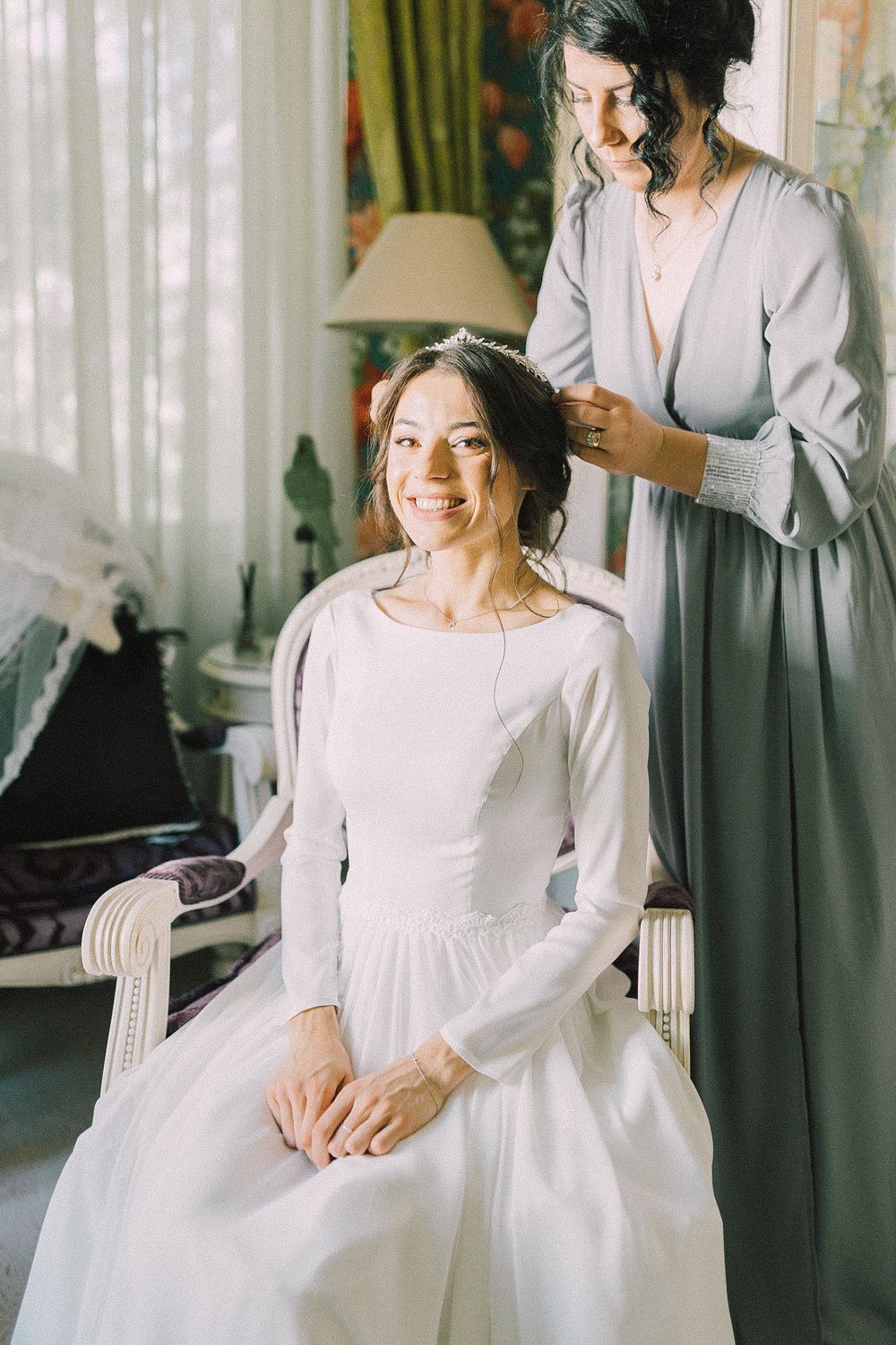 A smiling bride in a white gown sits while a bridesmaid in gray adjusts her hair in a bright, elegant room.